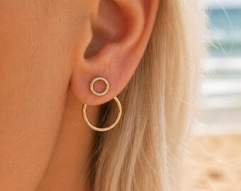 May include: Gold-colored double hoop earrings. The earrings feature a small, circular stud and a larger, open circle that dangles below. The earrings are shown on a person's ear, with blonde hair visible in the background. The earrings are a simple, elegant design.