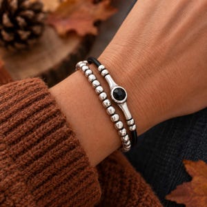 Könnte beinhalten: Ein zweireihiges Armband bestehend aus einer Reihe kleiner, polierter silberfarbener Metallperlen und einer zweiten Reihe aus einer glatten schwarzen Kordel, akzentuiert durch ein silberfarbenes Metallelement mit einem schwarzen Stein.