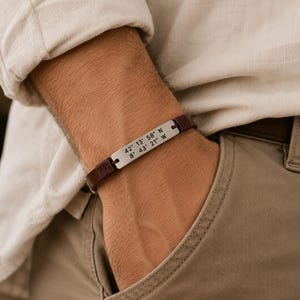 May include: A dark brown leather bracelet featuring a rectangular silver-toned metal plate engraved with the coordinates 42° 13' 58" N and 8° 43' 21" W. The band is secured with dark brown thread wrapping at the connection points to the plate.