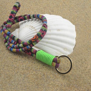 Può includere: Un cordino colorato con un motivo intrecciato nei toni del rosa, verde, blu e arancione. Il cordino ha una sezione verde brillante e un anello color oro. L'articolo è appoggiato su una superficie sabbiosa accanto a una conchiglia bianca.