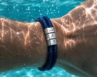 May include: A navy blue rope bracelet with three silver cylindrical beads. Each bead features a different etched icon: swimming, cycling, and running. The bracelet is shown underwater, with the wearer's arm visible.