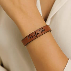 May include: A brown leather bracelet with the words "Me You Too" engraved in a cursive font. The bracelet is worn on a wrist, and the background is a white shirt.