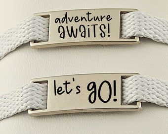 May include: Two silver metal shoe charms with gray fabric straps. The charms have black text that reads "adventure awaits!" and "let's go!"