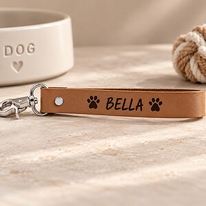 May include: A tan leather keychain featuring a silver metal lobster clasp and a silver rivet. The leather strap is engraved with the name BELLA centered between two black paw print icons, creating a personalized accessory for keys or bags.