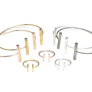 Op de afbeelding: Set van drie armbanden met staven in goud, rosé goud en zilver en twee bijpassende ringen. De armbanden en ringen hebben een eenvoudig, minimalistisch ontwerp met een enkele horizontale staaf.
