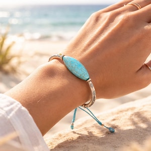 May include: A silver bracelet with a large, oval turquoise stone at the center. The bracelet has intricate floral designs on the sides and is secured with a blue cord. The background shows a beach with the ocean in the distance.