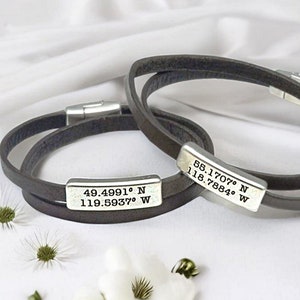 Puede incluir: Dos pulseras de cuero marrón con placas de metal plateado grabadas con coordenadas. Las coordenadas en la placa superior son 58.1707° N 118.7884° W. Las coordenadas en la placa inferior son 49.4991° N 119.5937° W.
