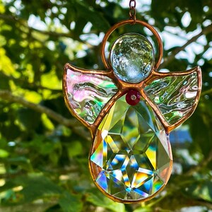 Peut inclure: Ornement en vitrail en forme d'ange, avec un corps en cristal facetté, des ailes irisées et une tête en verre transparent. L'ange est encadré de cuivre et possède une petite perle rouge au centre. Il est suspendu à une chaîne.