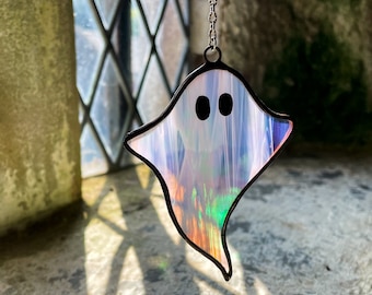 Puede incluir: Un atrapasoles de fantasma de vidriera con un contorno negro y una cadena para colgar. El fantasma tiene dos ojos negros y un acabado iridiscente brillante que refleja múltiples colores. El atrapasoles está colgado frente a una ventana.