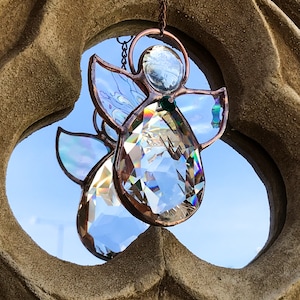 Pode incluir: Dois apanhadores de sol de anjo de cristal com molduras de metal cor de cobre e asas iridescentes pendurados contra um c&eacute;u azul. Os anjos est&atilde;o suspensos em frente a um arco de pedra texturizada castanho claro.
