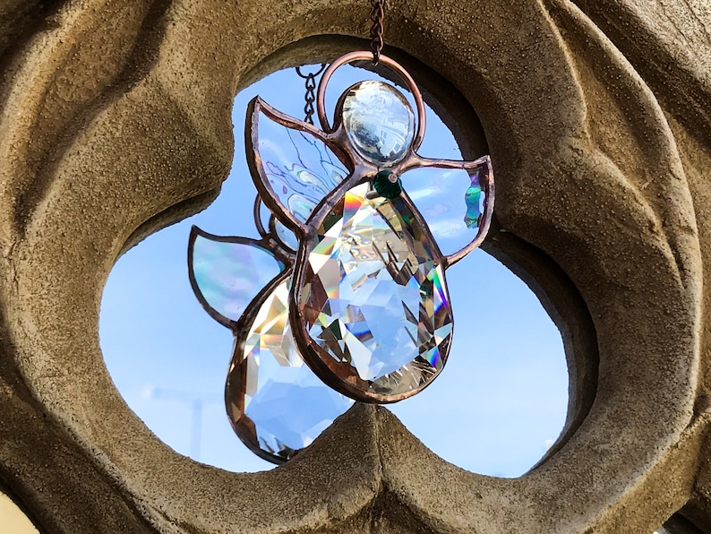 Pode incluir: Dois apanhadores de sol de anjo de cristal com molduras de metal cor de cobre e asas iridescentes pendurados contra um c&eacute;u azul. Os anjos est&atilde;o suspensos em frente a um arco de pedra texturizada castanho claro.
