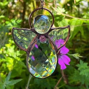 Pode incluir: Um apanhador de sol de anjo de vitral com cabe&ccedil;a e corpo de cristal transparente, asas iridescentes e uma gema rosa. O anjo &eacute; suspenso por uma corrente de cor de cobre, com um fundo verde desfocado e uma flor roxa.