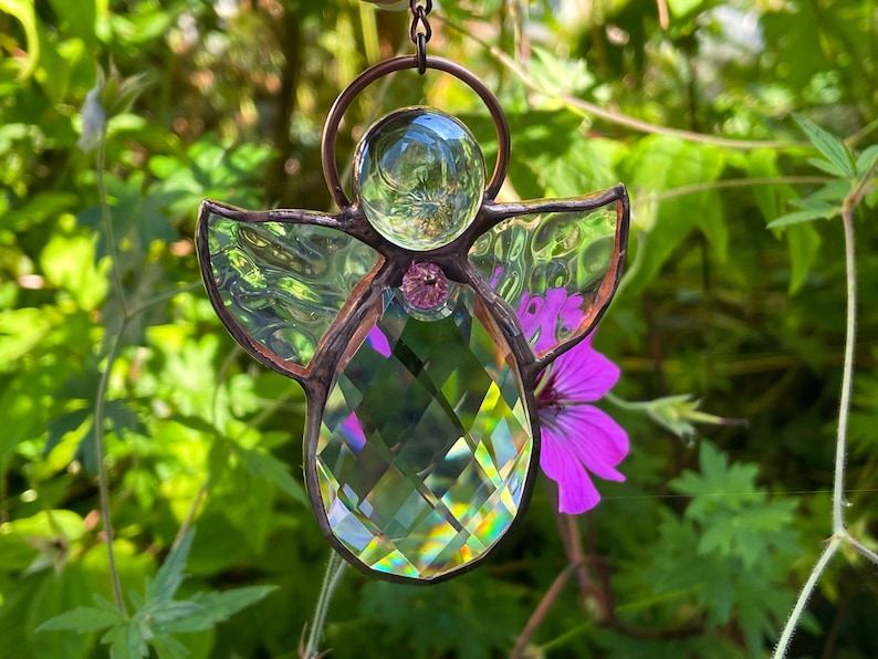 Pode incluir: Um apanhador de sol de anjo de vitral com cabe&ccedil;a e corpo de cristal transparente, asas iridescentes e uma gema rosa. O anjo &eacute; suspenso por uma corrente de cor de cobre, com um fundo verde desfocado e uma flor roxa.
