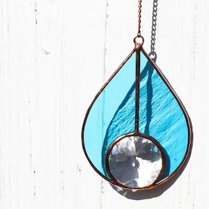 May include: A stained glass suncatcher in a teardrop shape, featuring a light blue panel and a clear, faceted crystal. The glass is framed with a copper-colored metal and hangs from a chain. The suncatcher is designed to catch and reflect light.