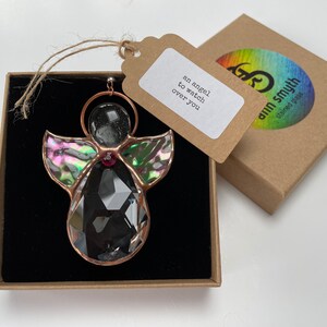 Pode incluir: Um enfeite de anjo de vitral com um corpo de cristal preto facetado, asas iridescentes e uma moldura de cobre. O enfeite est&aacute; em uma caixa marrom com uma etiqueta que diz "an angel to watch over you."