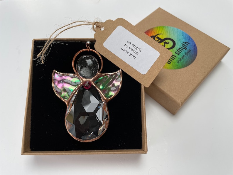 Pode incluir: Um enfeite de anjo de vitral com um corpo de cristal preto facetado, asas iridescentes e uma moldura de cobre. O enfeite est&aacute; em uma caixa marrom com uma etiqueta que diz "an angel to watch over you."