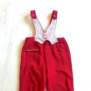 Puede incluir: Overol de beb&eacute; de pana rojo con un babero a rayas blancas y rojas. El babero tiene un borde festoneado y botones rosas. El overol tiene un bolsillo en la espalda y cierres de broche en los tobillos. Una prenda de beb&eacute; de estilo vintage.