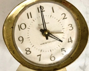 May include: Vintage Baby Ben Westclox alarm clock with a gold-tone bezel and a cream-colored face. The clock has black hands and numerals, with the words "Baby Ben" and "WESTCLOX" printed on the face. The clock is made in the USA.