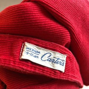 Puede incluir: Primer plano de una prenda de punto roja con una etiqueta que dice "MEDIUM (19-22 LBS.) Carters". La tela es de un rojo s&oacute;lido y vibrante con una textura acanalada. La etiqueta es blanca con texto azul.