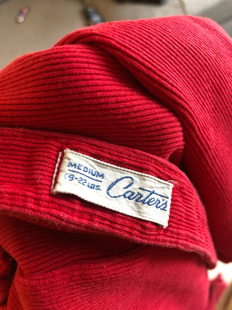 Puede incluir: Primer plano de una prenda de punto roja con una etiqueta que dice "MEDIUM (19-22 LBS.) Carters". La tela es de un rojo s&oacute;lido y vibrante con una textura acanalada. La etiqueta es blanca con texto azul.