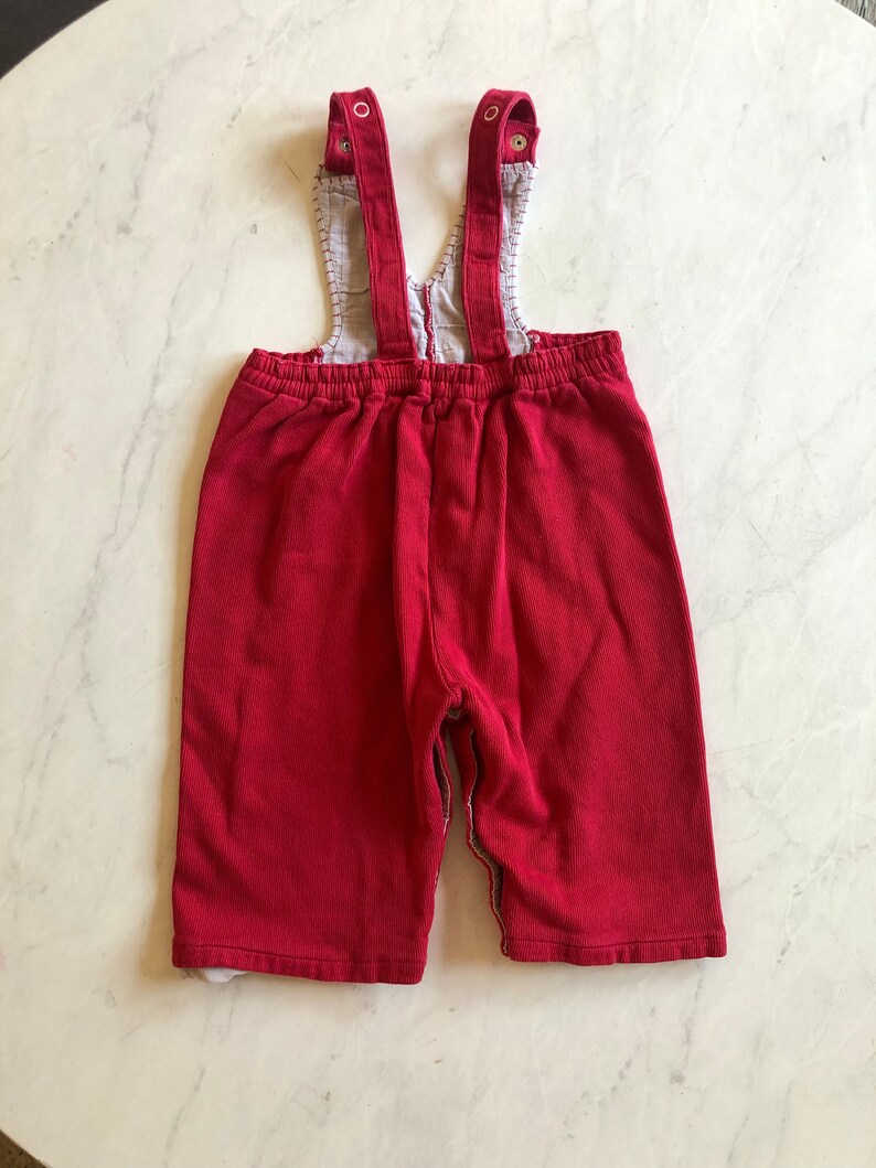 Puede incluir: Peto de beb&eacute; de pana rojo con tirantes ajustables. El peto tiene un forro estampado en contraste visible en el peto y los pu&ntilde;os. Los tirantes se abrochan con botones.