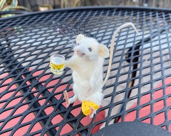 May include: A white mouse figurine holding a small glass of beer and a slice of cheese. The figurine is sitting on a black metal grate.