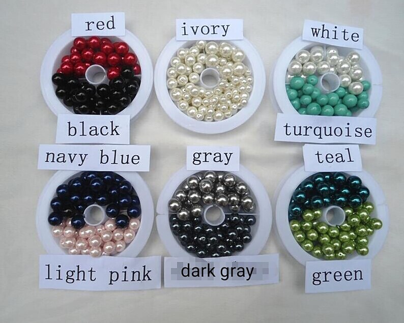 May include: Assortment of pearl beads in various colors, including red, ivory, white, turquoise, teal, navy blue, gray, light pink, dark gray, and green. The beads are displayed in small white containers with labels indicating their color.
