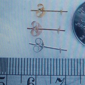 May include: Three pairs of earring posts in gold, rose gold, and silver tones. Each post has a butterfly-shaped backing and a small peg for securing an earring. A ruler is in the foreground.