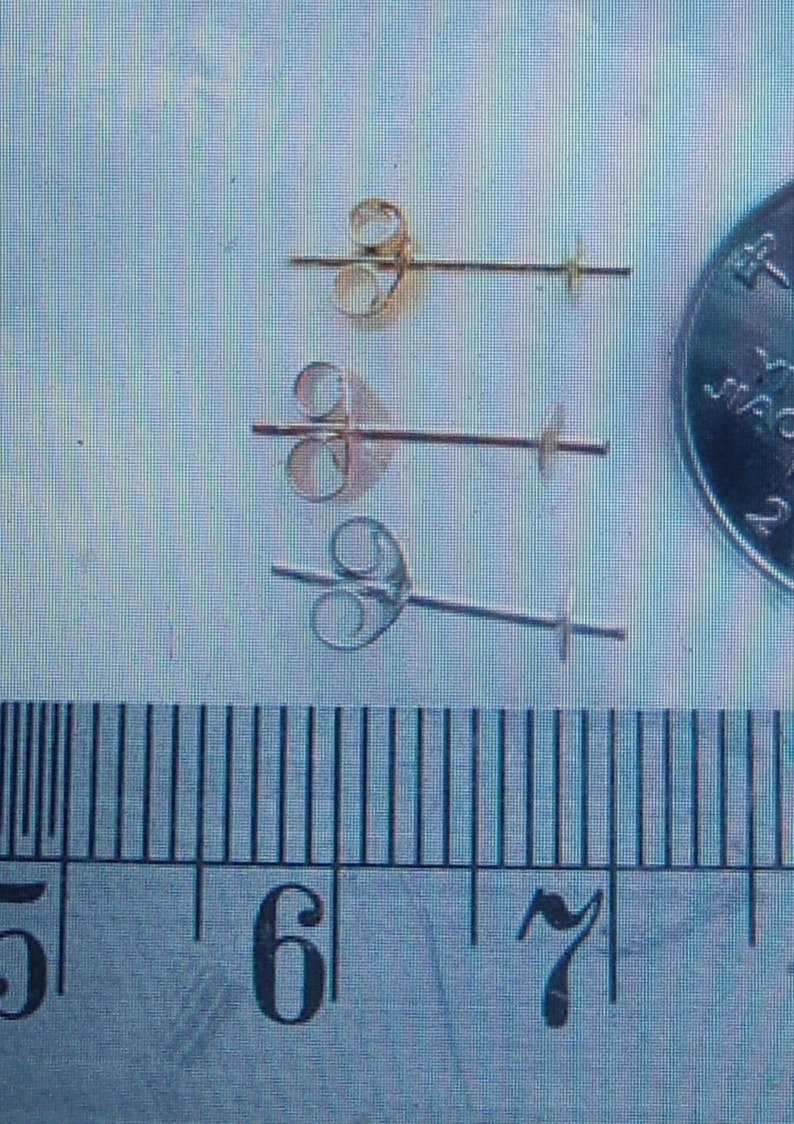 May include: Three pairs of earring posts in gold, rose gold, and silver tones. Each post has a butterfly-shaped backing and a small peg for securing an earring. A ruler is in the foreground.