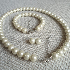 May include: A cream-colored pearl jewelry set, including a necklace, bracelet, and matching stud earrings. The necklace and bracelet feature round pearls strung together with a silver-tone chain clasp. The earrings are simple, round pearl studs.