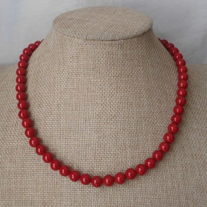 May include: A red beaded necklace on a fabric-covered mannequin bust. The necklace is made of round, smooth beads.