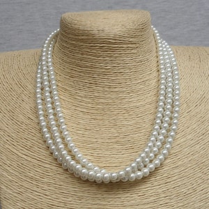 May include: A white faux pearl necklace with three strands of pearls. The necklace is displayed on a straw-colored mannequin bust.