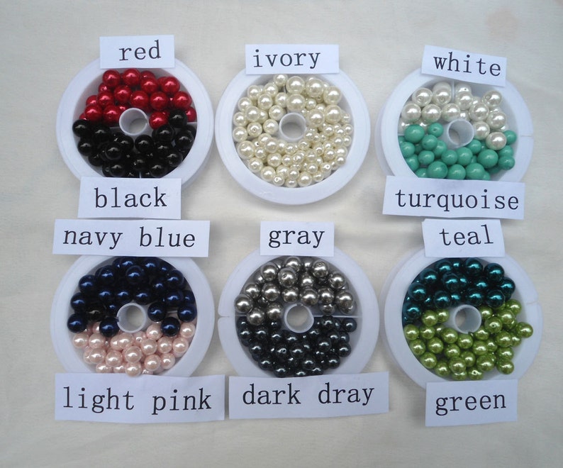 May include: An assortment of colorful beads in various shades, including red, black, ivory, white, turquoise, teal, navy blue, light pink, gray, dark gray, and green. Each color is labeled on a white tag. These beads are likely for jewelry making or crafting.