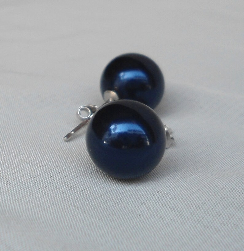 May include: A pair of dark blue, spherical stud earrings. The earrings have a glossy finish and are set on silver posts. The earrings are displayed on a light gray fabric background.