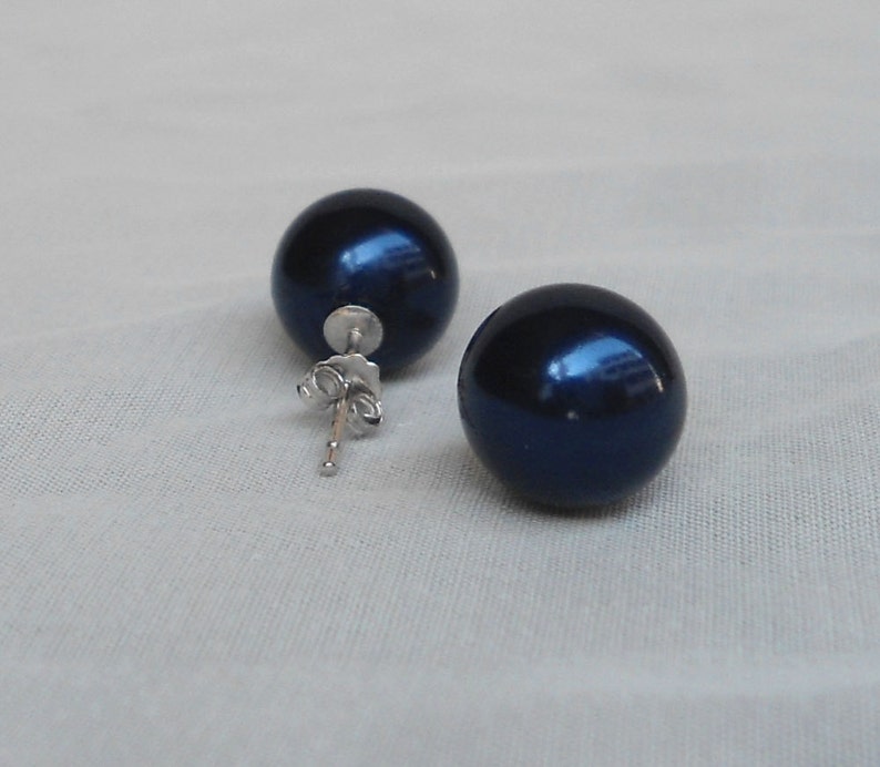 May include: A pair of dark blue, spherical stud earrings. The earrings have a glossy finish and are set on silver posts with butterfly clasps. The earrings are displayed on a light-colored, textured surface.