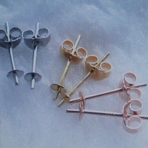 May include: An assortment of earring posts in silver, gold, and rose gold tones. The posts have a flat disc at the base for attaching earrings and a butterfly clutch backing. The earrings are displayed on a light blue background.