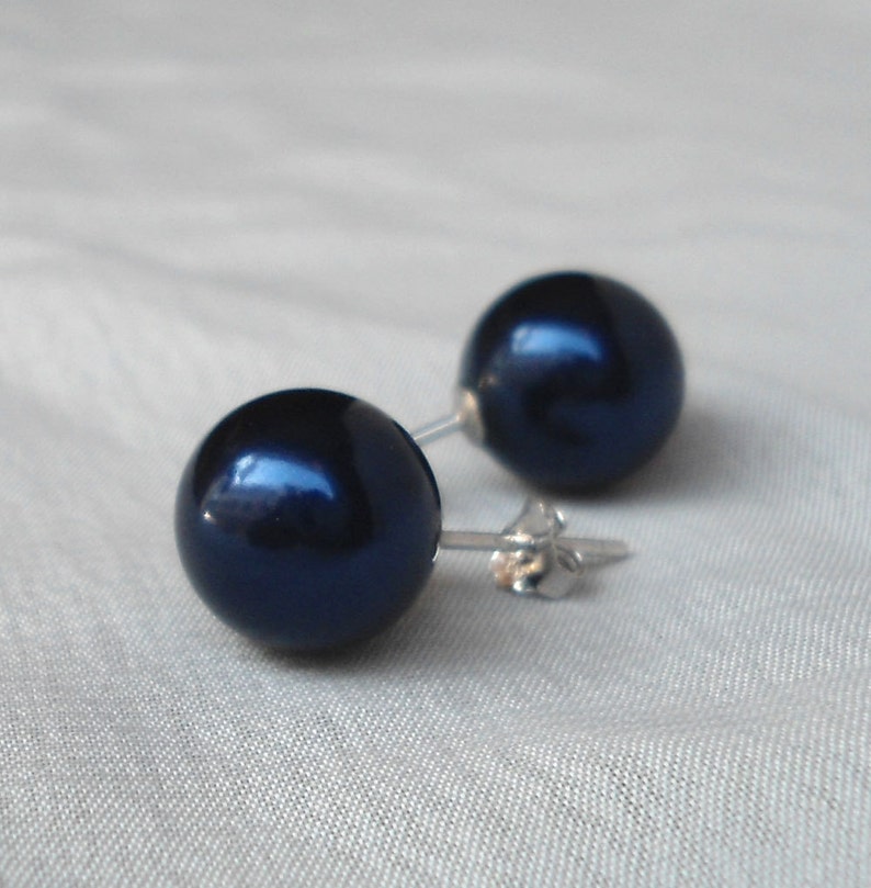 May include: A pair of dark navy blue stud earrings. Each earring features a large, round, glossy bead. The earrings have silver-colored posts and butterfly backs. The earrings are set against a light background.