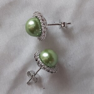 May include: A pair of silver earrings with green pearl accents. Each earring features a round green pearl surrounded by a circle of small clear stones.