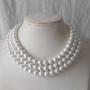 Puede incluir: Un collar de perlas blancas con tres hileras de perlas. El collar está en un busto de maniquí blanco.