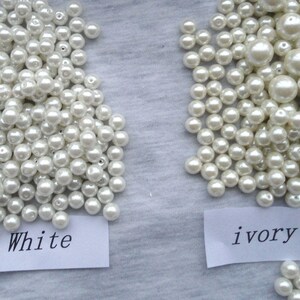 May include: Two piles of faux pearls, one labeled "White" and the other "ivory", are displayed on a gray surface. The pearls vary in size, with some larger than others. The ivory pearls have a warmer tone compared to the white pearls.