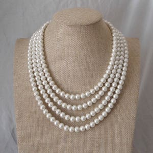 Può includere: Una collana di perle multifilo con quattro file di perle bianche. La collana è esposta su un manichino rivestito in tessuto.