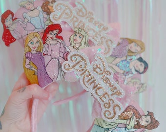 May include: A pink headband featuring multiple Disney Princess characters including Rapunzel, Ariel, and Aurora. The accessory includes a pink sequined bow with the text Disney Princess in gold glitter lettering, creating a layered, decorative design.