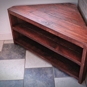 May include: A corner shelf made from reclaimed wood with two shelves. The shelf is stained a dark reddish brown color and has a rustic look.
