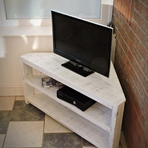 May include: White corner TV stand with two shelves. The stand is made of reclaimed wood and has a distressed finish. A flat screen TV is sitting on top of the stand.