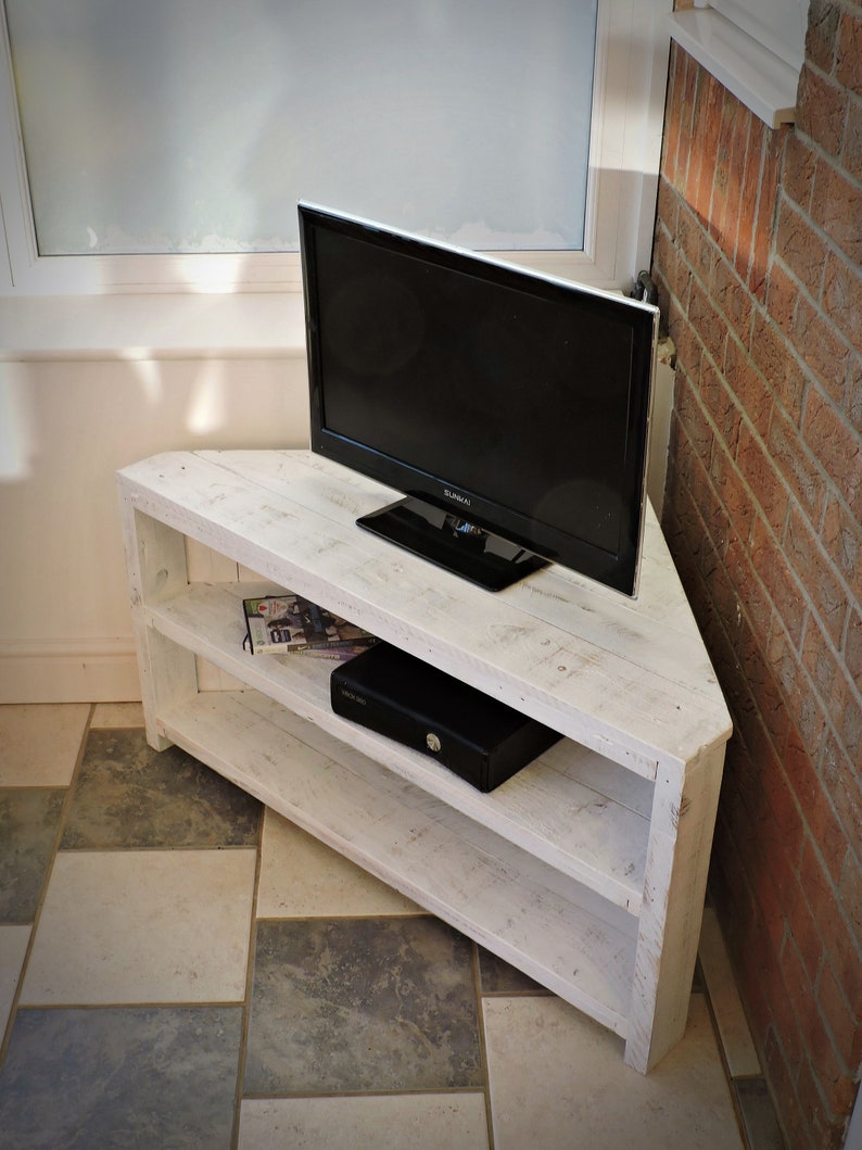 May include: White corner TV stand with two shelves. The stand is made of reclaimed wood and has a distressed finish. A flat screen TV is sitting on top of the stand.