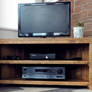 May include: A corner TV stand made of reclaimed wood with two shelves. The stand has a rustic brown finish and is holding a TV, a game console, and a stereo receiver.