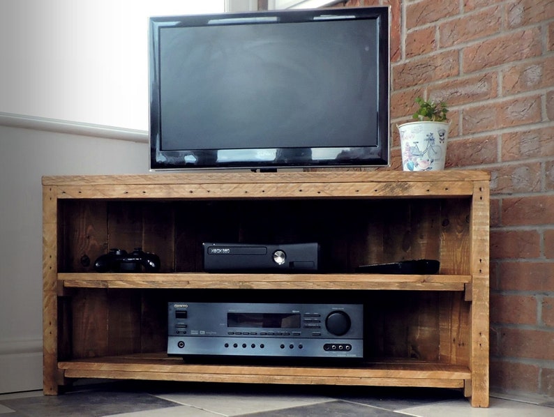 May include: A corner TV stand made of reclaimed wood with two shelves. The stand has a rustic brown finish and is holding a TV, a game console, and a stereo receiver.