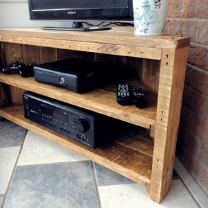May include: A corner TV stand made of reclaimed wood with two shelves. The stand is stained a light brown color and has a rustic look. It is holding a black gaming console, a black receiver, and two black remotes.