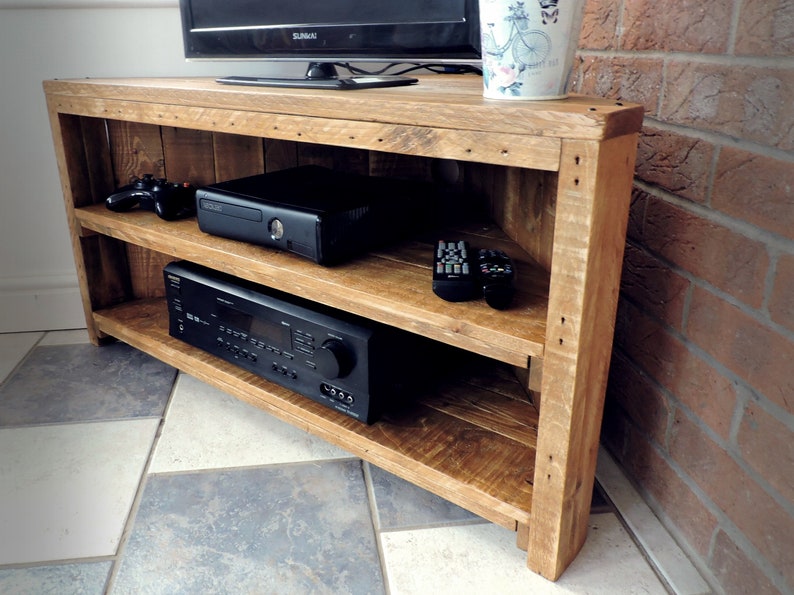 May include: A corner TV stand made of reclaimed wood with two shelves. The stand is stained a light brown color and has a rustic look. It is holding a black gaming console, a black receiver, and two black remotes.