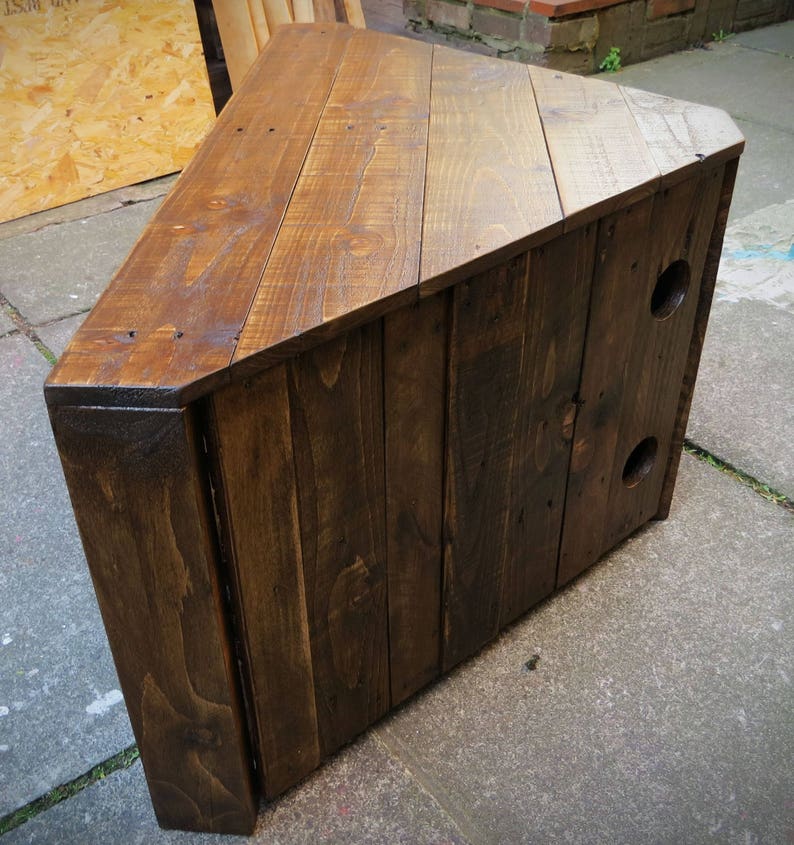 May include: A dark brown wooden corner TV stand with a triangular top and two holes on the side. The stand is made of reclaimed wood planks.